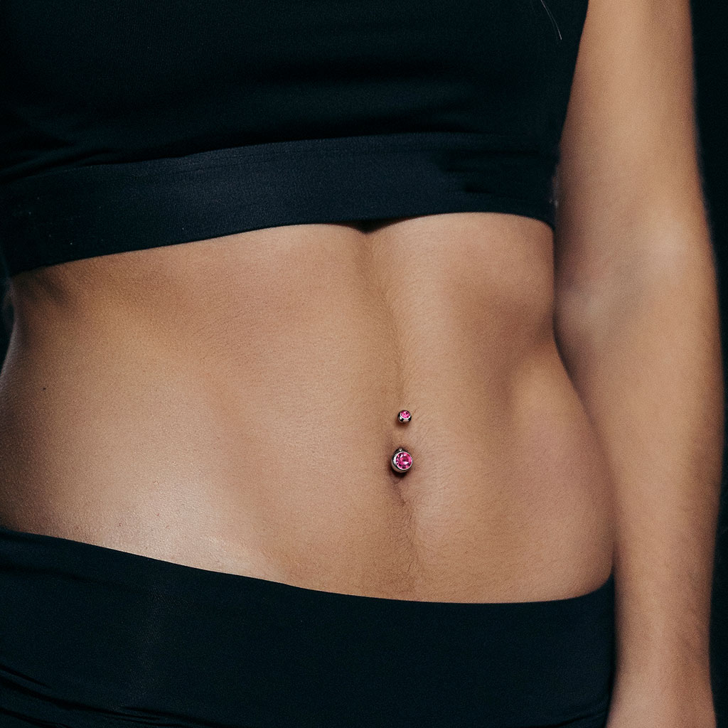 Woman with tan abs wearing black sports bra and black leggings with a popular pink fifth cue belly ring